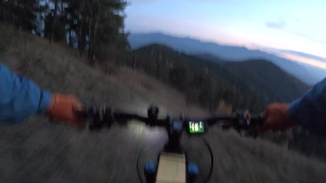 Chest view biker riding on e-bike along the edge of a steep cliff. Cyclist rides mountain bicycle evening field trail background hills covered green summer forests. Extreme enduro e-mtb ride lantern