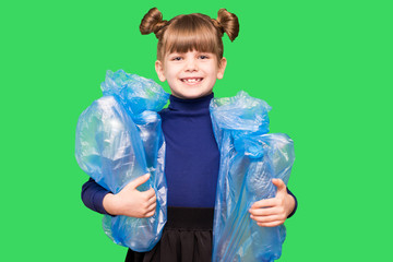 Positive little ecologist girl holding trash bags with plastic bottles isolated on green background