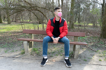 Portrait of brunette man in a surgical bandage sitting in the park on the bench, coronavirus, illness, infection, quarantine, medical mask