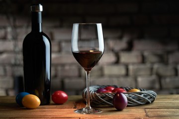 Easter decorations, colorful eggs, a glass of red wine with a bottle on table at home dark warm living room, brick wall in background. Copy space for text.