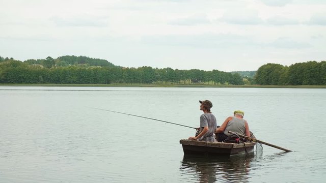 Old angler with a grandson sitting in the boat on the lake with a rod while fishing. Outside.