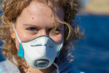 Close-up photo of young beatiful girl in medical protection face mask. Woman with blue eyes and curly blonde hair. Blurry background