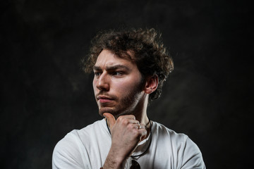 Fototapeta premium Curly and hansome caucasian man standing in a dark studio on a grey background, wearing casual white shirt looking thoughtful