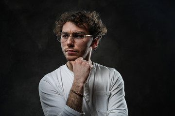 Close-up portrait of a young handsome male model posing on a grey background in a dark studio, wearing white casual shirt and looking serious