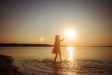 Silhouette of a girl running on water against the background of an evening sunset. Water splashes in all directions. Carefree happy baby fun.