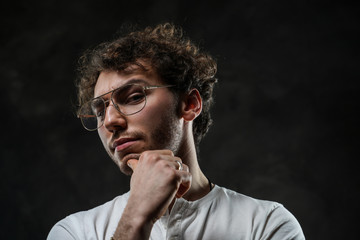 Close-up portrait of a young handsome male model posing on a grey background in a dark studio, wearing white casual shirt and glasses, looking interested