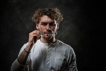 Curly and serious caucasian man standing in a dark studio on a grey background, wearing casual white shirt and glasses.