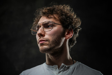 Obraz premium Curly and serious caucasian man standing in a dark studio on a grey background, wearing casual white shirt and glasses.