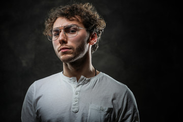 Thoughtful young student posing in a dark studio on a grey background, looking focused while wearing casual white shirt and glasses.