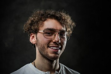 Fototapeta premium Close-up portrait of a young handsome male model posing on a grey background in a dark studio, wearing white casual shirt and glasses, looking joyful
