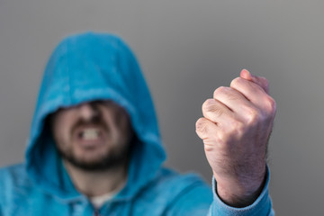 Angry man with unshaven face dressed in blue hoodie showing a fist. Selective focus. Fist in focus,...