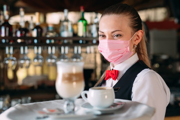 A female Waiter of European appearance in a medical mask serves Latte coffee.