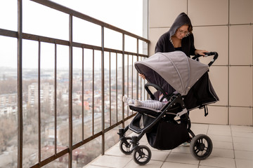 A young woman with a stroller and a child stands on the balcony of the apartment and enjoy the fresh air. Sleep in the air. Self-isolation and quarantine.