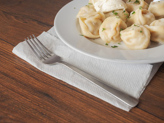 Traditional russian large dumplings with sour cream on a white plate and wooden table, and a metal fork.