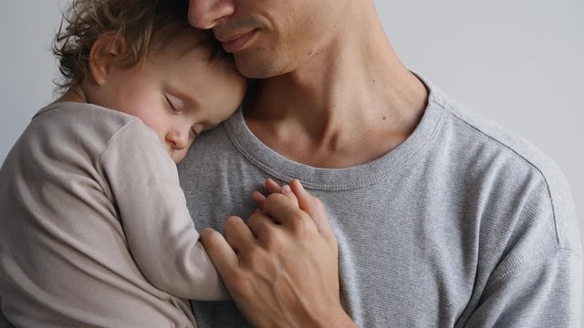 Close up portrait of a father and his daughter who sleeps in his arms. Father holds daughter's hand and smiles