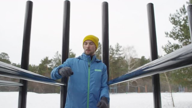 Medium shot with low angle of active man in blue windbreaker and yellow beanie hat walking up to workout bard at outdoor gym and doing dips while training in winter