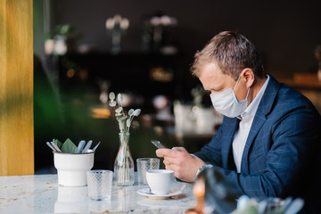 Male entrepreneur wears medical mask during cornovirus outbreak, sits at cafe, waits for business partner, drinks cup of coffee, checks newsfeed, connected to wireless internet. Health care concept