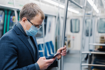Naklejka na ściany i meble Serious man commutes to work in underground wears virus protective mask being always in touch with modern cellular poses in empty subway or metro. Distance concept. Public transport during coronavirus