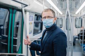 Fototapeta na wymiar Shot of young man wears spectacles and protective face mask to prevent spreading coronavirus disease or flu epidemic in public transport, poses at empty subway carriage. Public health solution