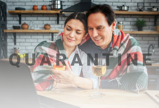 Cozy picture of couple sitting together at table in kitchen. They look at laptop and smile. Their shoulders covered with blanket. Glasses of wine stand at table.