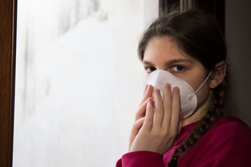 Young girl in home isolation, during quarantine is wearing protective face mask for stave off preading of disease coronavirus, near the window