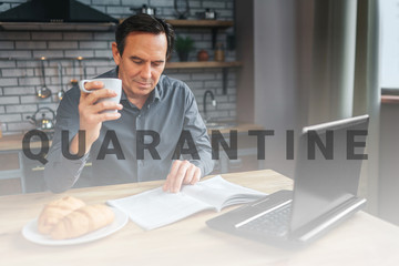 Adult businessman sit at table in kitchen anr read journal. He hold white cup and look down. Laptop and plate with croissans on table.