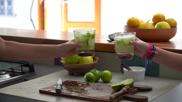 Three friends hands celebrating and toasting glasses with Brazilian Caipirinha drink, made with cacha&ccedil;a and lemon at home during sunny summer day. Friendship, happiness, celebration concept.