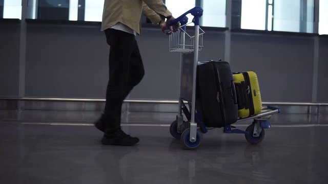 Unrecognizable male passenger with luggage trolley in the international airport. Walkning in the hurry, side view