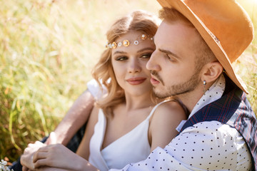 A young man in a hat and a beautiful girl with a make-up and hairstyle, hugging sitting in a field in the grass on a Sunny day