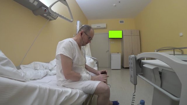 The patient listens to music from a smartphone in headphones sitting on the bed then lies down. A man in a white hospital shirt in a single room surrounded by functional furniture.