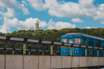Older metro train in Kiev is running over the bridge over Dnieper river towards Hydropark and Dnipro district on a warm summer day in Ukraine. Laura churches complex is seen in the background, a Kiev 