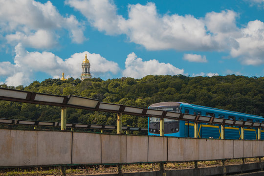 Newer metro train in Kiev is running over the bridge over Dnieper river towards Hydropark and Dnipro district on a warm summer day in Ukraine. Laura churches complex is seen in the background, a Kiev 