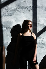 Girl in black sportswear standing near a grey stone-textured walls and looks out the window. Contrast the photo of the girl the light.