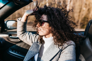 Fototapeta na wymiar Curly beautiful woman in a convertible car on a sunny day in sunglasses relaxes and feels freedom to ride.