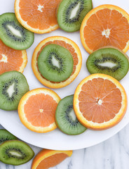 Close up of sliced oranges and kiwi on a white plate on a marble background.