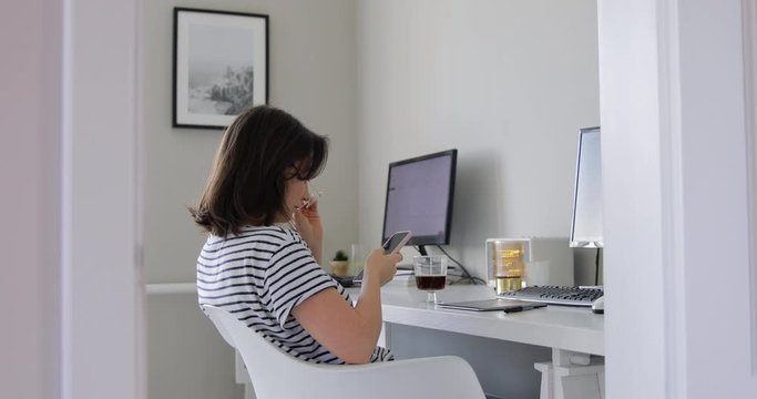 A portrait of a pretty young girl with brown hair wearing eyeglasses web surfing in a living room office with computers. Artistic indoor. Busy businesswoman taping and texting on her smartphone