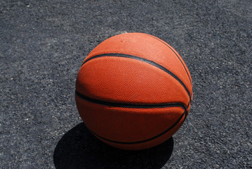 A single basketball sits on a dark pavement court in the sun. The close up of the ball shows it is orange, round, and leather with dark black stripes. The ball sits on an outdoor court ground.