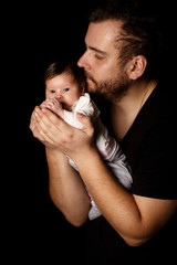 a bearded father in a black t-shirt holds a newborn baby. Dad kisses and hugs daughter or son on a black background