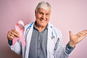 Senior handsome hoary doctor man wearing stethoscope holding pink cancer ribbon very happy and excited, winner expression celebrating victory screaming with big smile and raised hands