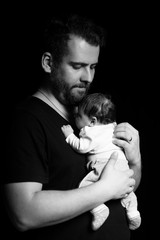 a bearded father in a black t-shirt holds a newborn baby. Dad kisses and hugs daughter or son on a black background