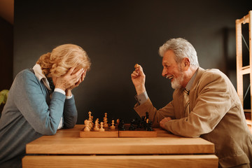 Close photo of elderly couple playing chess at home. Woman is not happy because man is winning.