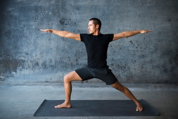 Man practicing yoga in front of a dark background. Pose is warrior II, Virabhadrasana. 