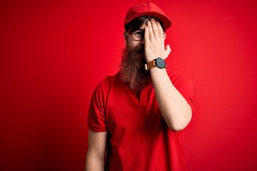 Young handsome delivery man wearing glasses and red cap over isolated background covering one eye with hand, confident smile on face and surprise emotion.