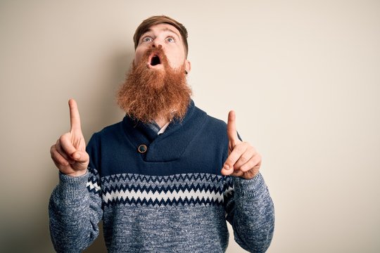Handsome Irish redhead man with beard wearing winter sweater over isolated background amazed and surprised looking up and pointing with fingers and raised arms.