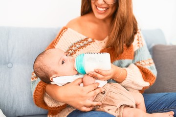 Young beautiful woman and her baby on the sofa at home. Newborn and mother relaxing and resting comfortable drinking milk using feeding bottle