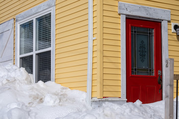 A yellow exterior wooden house with a red door and white trim windows with a drift of snow in front. The house has narrow clapboard on the outside with a metal red door and closed glass window.   - Powered by Adobe