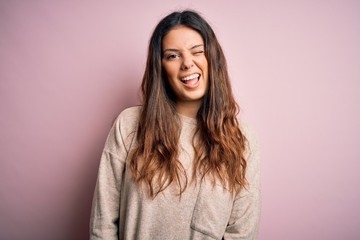 Young beautiful brunette woman wearing casual sweater standing over pink background winking looking at the camera with sexy expression, cheerful and happy face.