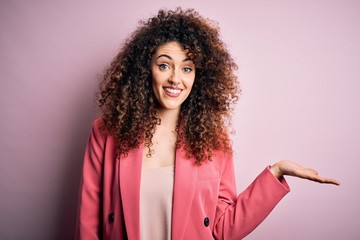 Young beautiful businesswoman with curly hair and piercing wearing elegant jacket smiling cheerful presenting and pointing with palm of hand looking at the camera.