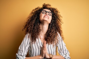 Young beautiful woman with curly hair and piercing wearing striped shirt and glasses begging and...