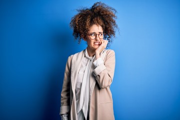 Fototapeta na wymiar Young beautiful businesswoman with curly hair and piercing wearing jacket and glasses looking stressed and nervous with hands on mouth biting nails. Anxiety problem.